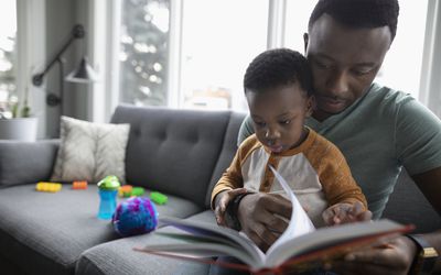 Father and toddler son reading book on living room sofa