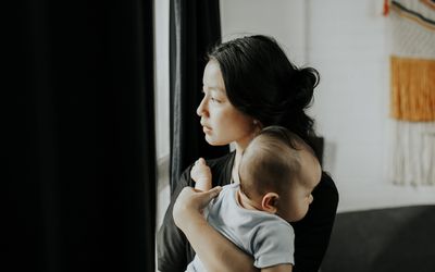 A young mother looks out the window into the light while holding her young infant to her chest