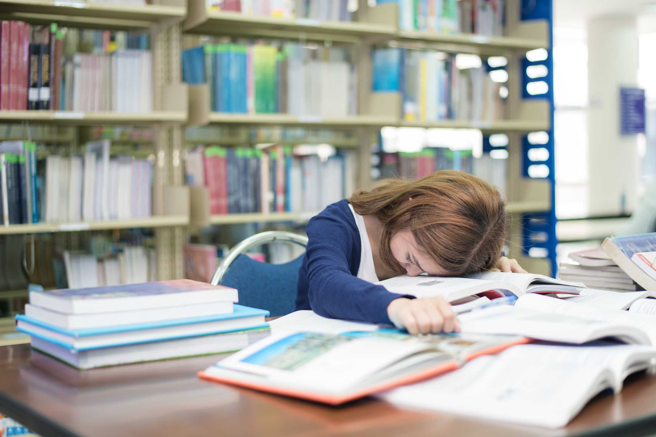 疲惫的学生或年轻女子与许多书睡觉，而阅读书籍准备考试在图书馆在大学。人、教育、会议、考试和学校概念