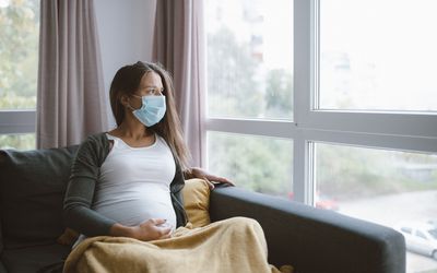 A pregnant person sits on a couch with a face mask on and a blanket while staring out the window.