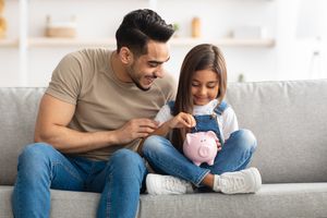 可爱的微笑小女孩把硬币放在粉红色的存钱罐里，坐在家里的沙发上和爸爸，男人教他的女儿如何投资