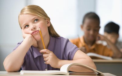 Girl (6-7) sitting in classroom with head in hands, students in background.