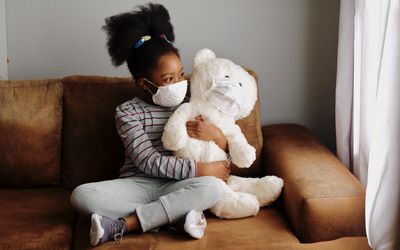 Young girl wearing a mask, holding teddy bear wearing a mask on a couch