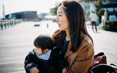 年轻的亚洲单身母亲抱着可爱的小女儿，坐在户外码头边沉思