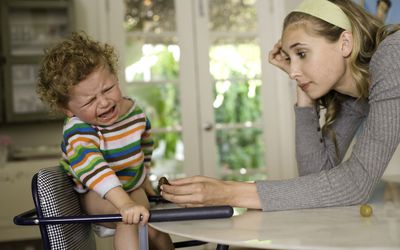 蹒跚学步的孩子在高椅子上挣扎和哭泣，而排水的女人试图在餐桌上提供食物。