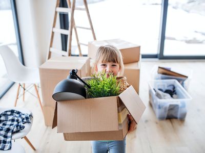 Little girl carrying a moving box holding a lamp and plant.