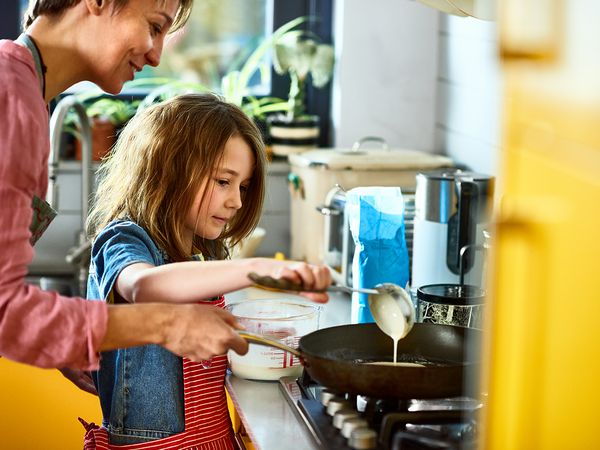 Girl pouring pancake batter in frying pan, with mother