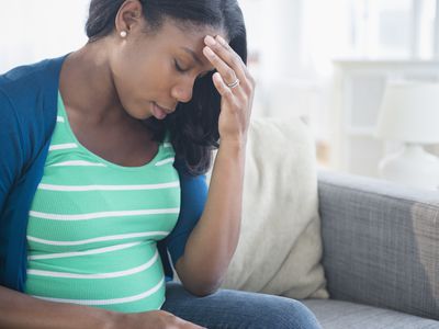 Anxious Black pregnant woman rubbing forehead on sofa