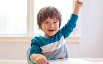 Toddler smiling and raising his hand at a desk while holding pen with his other hand