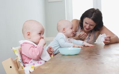 一对双胞胎女婴坐在桌旁，一个用勺子喂，另一个嚼着她的拳头