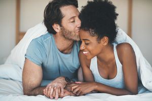 一名深情的中年男子在家里的床上放松时亲吻妻子的额头。＂width=