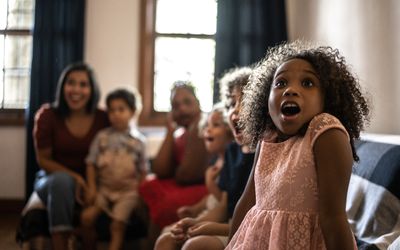 A young black girl in the foreground happily gasps at something on the TV, out of frame, with her family.