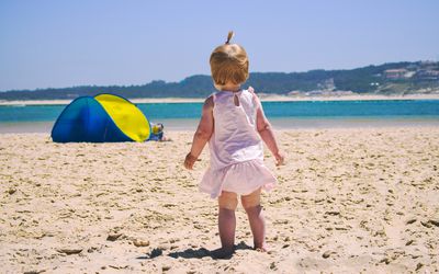 Rear View Of Baby Girl Standing On Sand At Beach Against Sky
