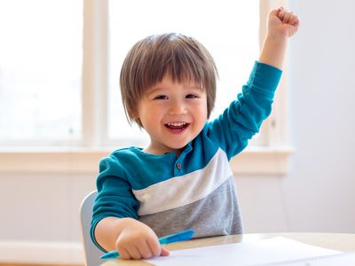 Toddler smiling and raising his hand at a desk while holding pen with his other hand