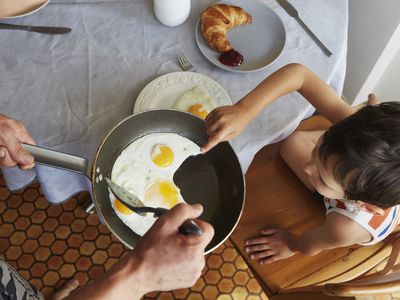 Parent offering child frying pan of sunnyside up eggs at breakfast table