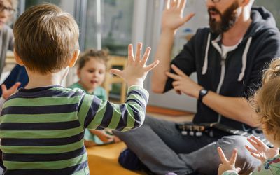 Child raising hand while singing with others and teacher in preschool