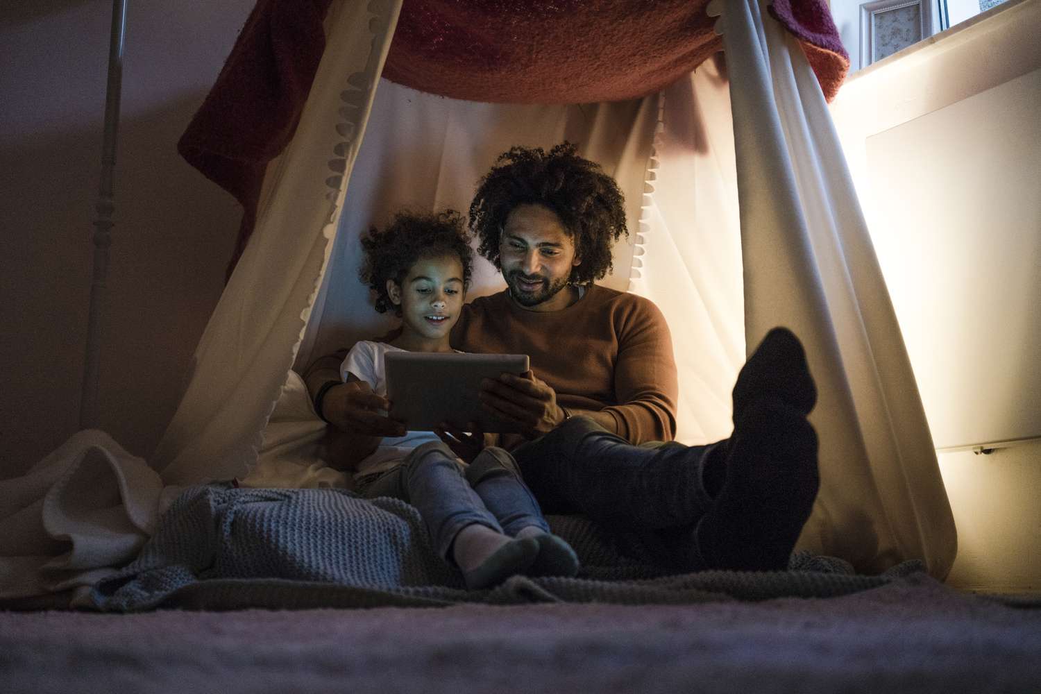 Father and daughter sitting in dark childrens room, looking at digital tablet