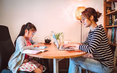 年轻的亚洲妈妈在家里用笔记本电脑工作，而她的小女儿在家里的工作空间里用数字平板电脑阅读电子学习资源。