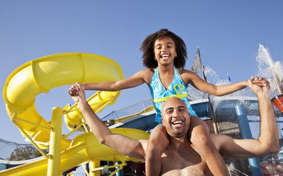 在水上公园里，一个穿着蓝色比基尼的小女孩微笑着坐在她父亲的肩膀上。
