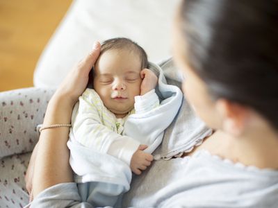 Baby smiling in sleep on mother's lap. Nestled in a blue shawl. Mother is white with palm around baby's right ear. Mother wearing grey leggings, spotted with a red print