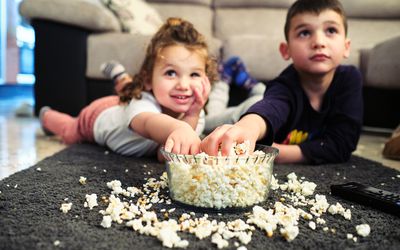 两个小孩趴着看电影，一边啃着一碗溢出来的爆米花。