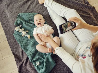 A mother takes a video of a baby with a smartphone. The baby is laying on a green blanket and is wearing a white onesie. There are toys scattered around.