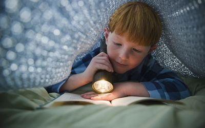 Young boy reading a book with a flashlight under the covers