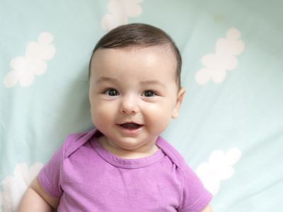 Baby boy in purple onesie looking at the camera and smiling lying against cloud sheets