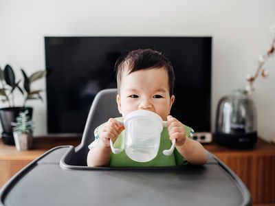 An Asian infant holds a clear sippy cup with two hands in a high chair.