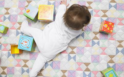 Overhead view of a baby in a white outfit playing with blocks on a colorful quilt.