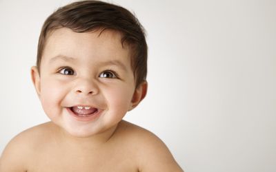 Baby boy with brown hair brown eyes smiling with two teeth showing, looking past the camera with no shirt on