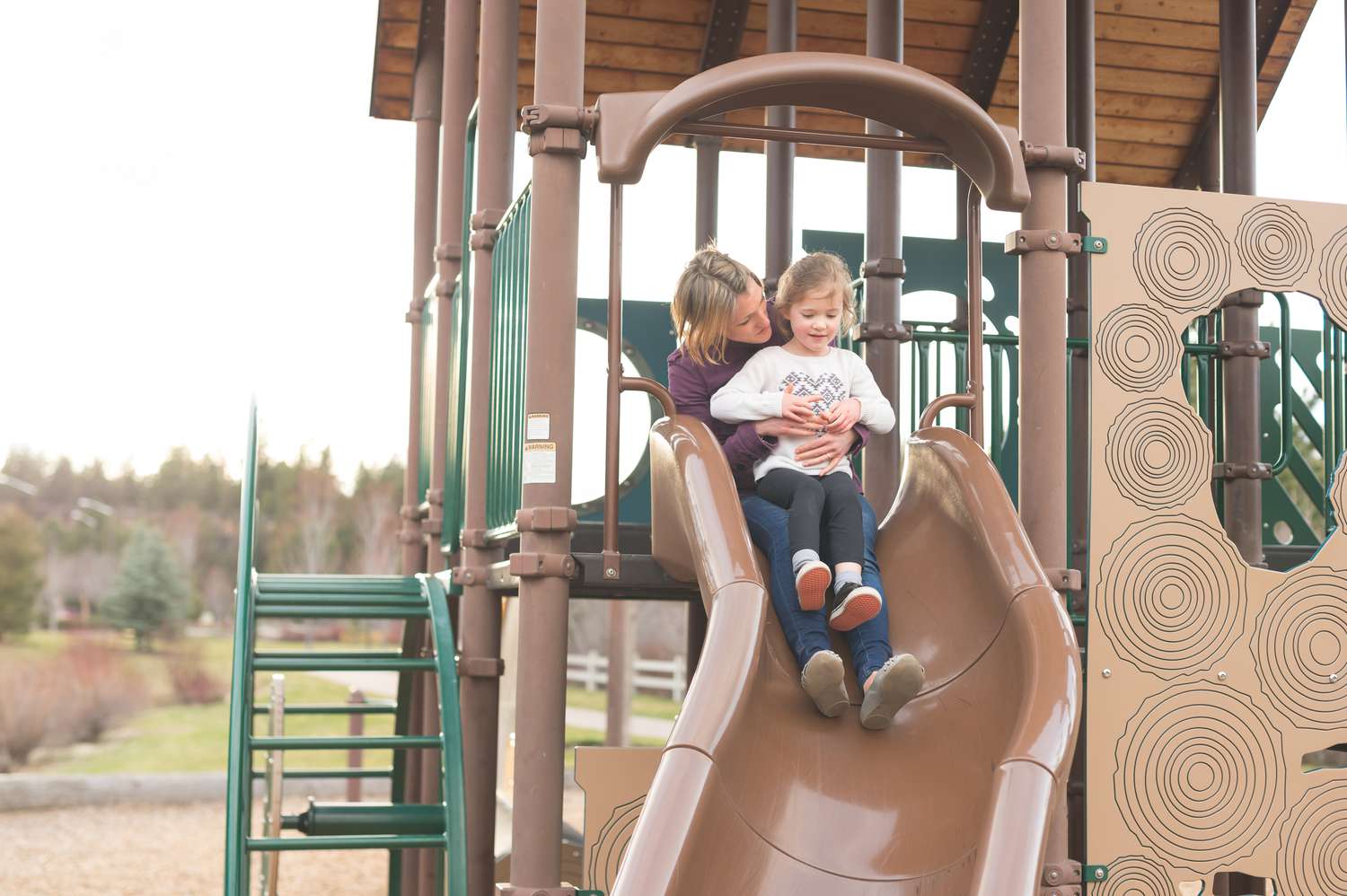 Little girl sits on her mother's lap going down a playground slide