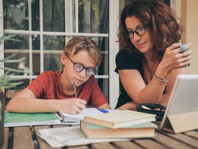 Young student doing homework at home with school books newspaper, digital pad helped by his mother. Mum control, help and teaching his son. Education, family, lifestyle and homeschooling concept.