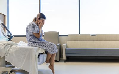 A woman in a hospital gown sits on the edge of a hospital bed, head in hands.