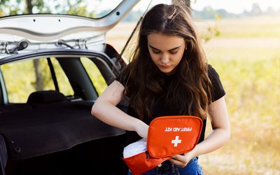 Young attractive girl stands near car with open back door and emergency lights is on, tries to find something in first aid kit