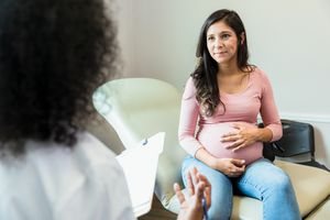 a pregnant person touches her abdomen as she listens to a doctor. The doctor gestures as she holds the patient's medical records.