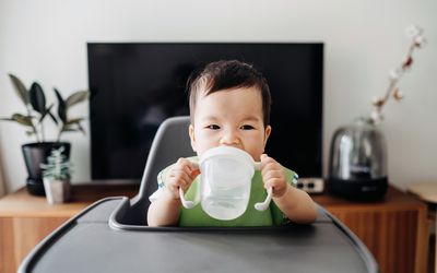 一个亚洲婴儿双手捧着一个透明的吸管杯，放在高脚椅上。