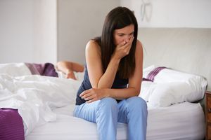 Woman with hand over mouth sitting on edge of bed looking sick