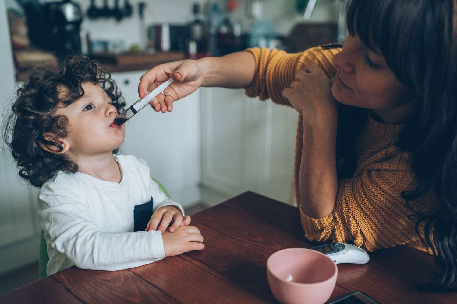 A mother gives her toddler liquid medicine via syringe while sitting at the kitchen table together.