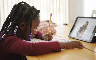 A girl sits at a table doing online school writing with a pencil.