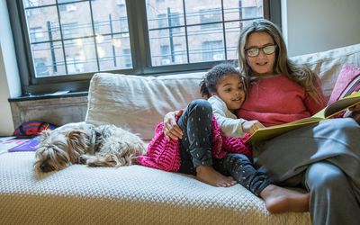 小女孩和她的妈妈一起在沙发上看书，旁边有狗＂width=