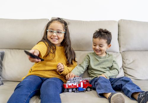 Sister and brother watching TV and playing with a firetruck on the couch.