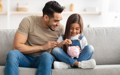 可爱微笑的小女孩把硬币放进粉红色的小猪存钱罐里的肖像，和爸爸坐在家里的沙发上，男人教他的女儿如何投资