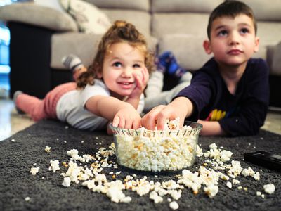 Two little kids laying on their bellies watching a movie while digging into an overspilling bowl of popcorn.
