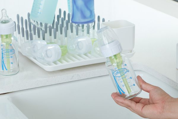 Hand holding Dr. Brown's Natural Flow Options+ Narrow Glass Baby Bottle over a sink next to a dish drying rack with other baby bottles