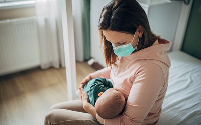 Mom wearing mask breastfeeds newborn in hospital room