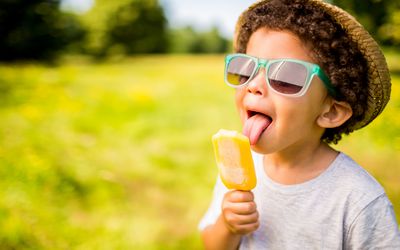 Little boy outside in a hat and sunglasses eating a popsicle.