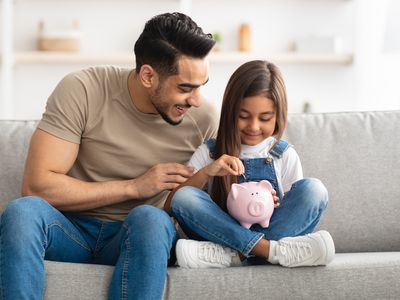 Portrait of cute smiling little girl putting coin in pink piggy bank, sitting with dad on the couch at home, man teaching his daughter how to invest