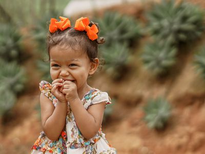 Toddler giggling with orange hairbows outside with plants in the background