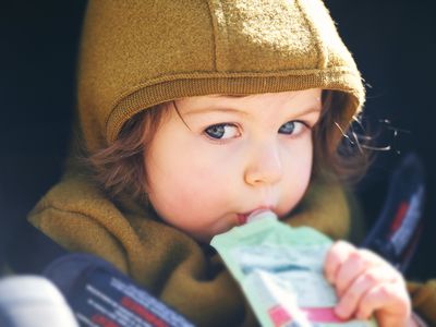 Close up portrait of sweet toddler kid eating fruit puree from plastic doy pack, sitting in stroller, outdoor snack time